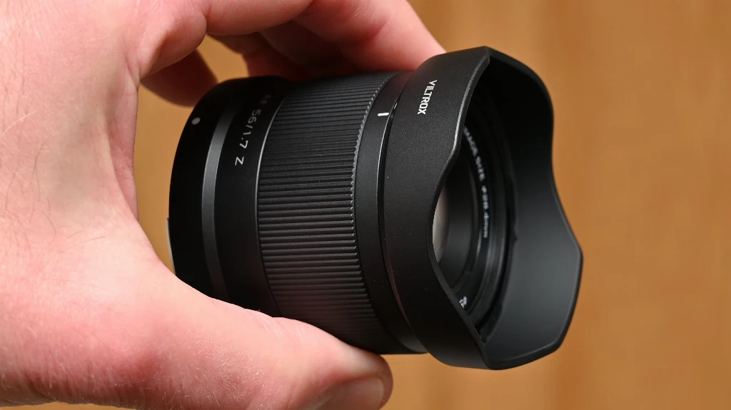 Объектив Viltrox AF 56mm f/1.7 Air крупным планом в руке взрослого человека с надетой пластиковой лепестковой блендой на коричневом фоне