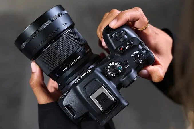 Объектив Sigma 56mm f/1.4 DC DN Contemporary с блендой LH582-01 на корпусе Canon EOS R в руках фотографа на фоне бетонной стены